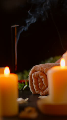 Vertical-Video-Shot-Of-Person-Putting-Down-Towel-With-Lit-Candles-With-Scattered-Petals-Incense-Stick-Against-Dark-Background-As-Part-Of-Relaxing-Spa-Day-Decor